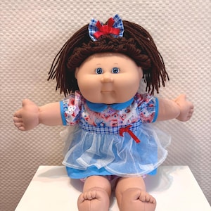 May include: A doll with brown yarn hair, blue eyes, and a round face. It wears a blue and white chequered dress with a floral print bodice, a blue collar, and a white tulle overlay. A small blue and white chequered bow sits atop its head.