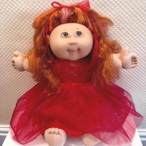 May include: A Cabbage Patch Kids doll with light skin, round cheeks, and a dimple on the left cheek. The doll has long, wavy, reddish-orange hair with a red ribbon bow and wears a sleeveless red dress featuring a glittery tulle skirt overlay.