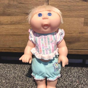 May include: A vintage Cabbage Patch Kids doll with blonde hair and blue eyes. The doll is wearing a pastel striped top with ruffled sleeves and light blue shorts with a graphic. The doll is standing on a patterned surface.