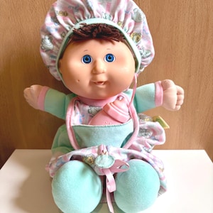 May include: A doll with brown yarn hair and bright blue eyes, wearing a light mint green and pink patterned outfit with a matching bonnet. The doll features a soft fabric body and includes a small pink plastic baby bottle tucked into a pocket.