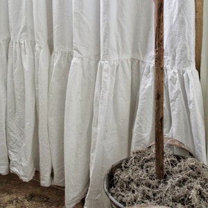Puede incluir: Cortina de ducha de algod&oacute;n blanco con un dobladillo con volantes. La cortina est&aacute; colgada en un ba&ntilde;o y tiene un palo de madera r&uacute;stico delante.