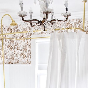 Puede incluir: Una barra de cortina de ducha dorada con cortinas blancas colgando de ella. La barra est&aacute; instalada en un ba&ntilde;o con papel tapiz floral y una l&aacute;mpara de ara&ntilde;a de cristal.