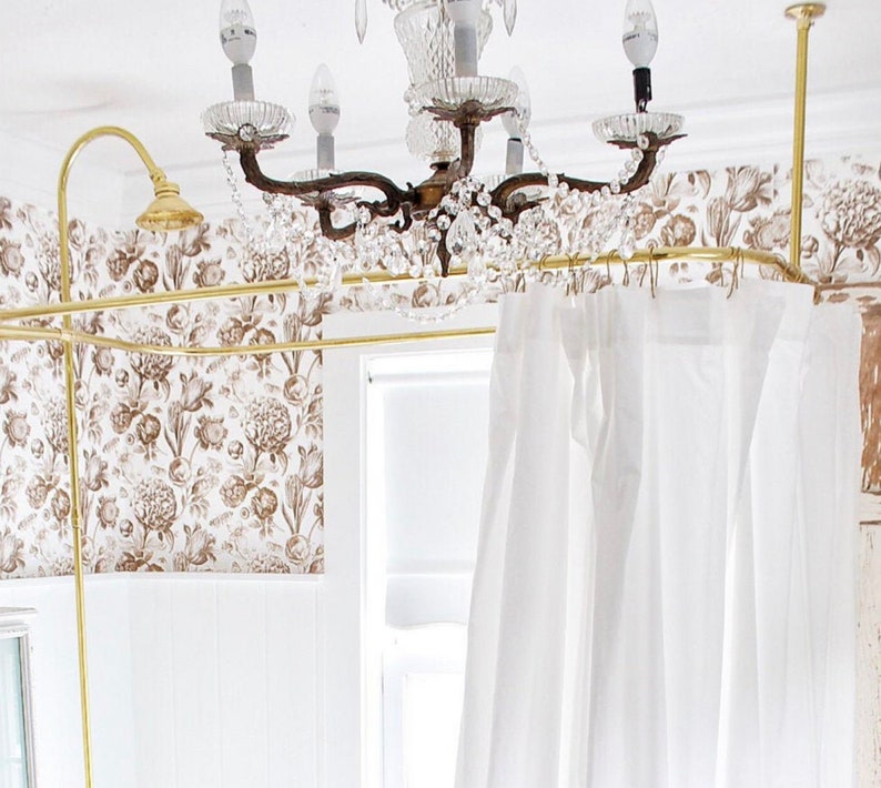 Puede incluir: Una barra de cortina de ducha dorada con cortinas blancas colgando de ella. La barra est&aacute; instalada en un ba&ntilde;o con papel tapiz floral y una l&aacute;mpara de ara&ntilde;a de cristal.