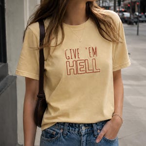 以下が含まれることがあります： マスタードイエローのTシャツ。赤い文字で「GIVE 'EM HELL」と書かれています。クルーネックのTシャツは、青いジーンズと組み合わせています。モデルは茶色のショルダーバッグを着用しています。生地は綿のようです。