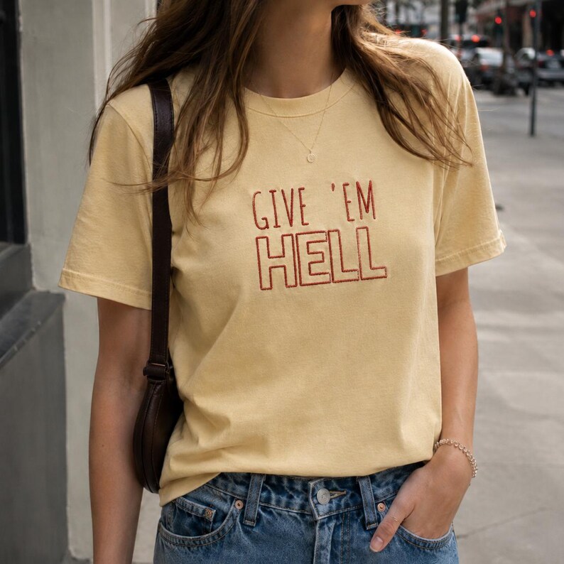 以下が含まれることがあります： マスタードイエローのTシャツ。赤い文字で「GIVE 'EM HELL」と書かれています。クルーネックのTシャツは、青いジーンズと組み合わせています。モデルは茶色のショルダーバッグを着用しています。生地は綿のようです。