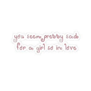 Op de afbeelding: Een gestanste sticker met de tekst you seem pretty sad for a girl so in love in een oudroze cursief lettertype met decoratieve krullen aan de letters. De sticker heeft een witte rand die de vorm van de tekst volgt.