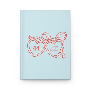 May include: A light blue hardcover journal featuring a coral-colored graphic of two connected heart-shaped lockets tied with a ribbon. The left heart contains the number 44, and the right heart displays the text Garrett Graham in script.