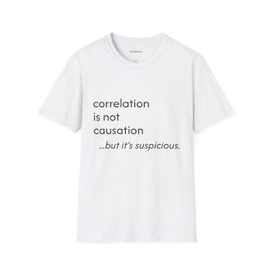 May include: White t-shirt with the text "correlation is not causation...but it's suspicious." The shirt has a crew neck and short sleeves. The text is in a simple, sans-serif font.