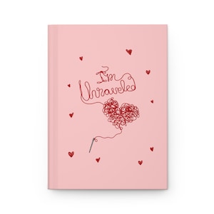 May include: A light pink hardcover journal featuring a red graphic of a tangled yarn heart shaped like a ball of thread with a needle attached. The text "I'm Unraveled" is written in a red cursive script above the heart, surrounded by small red hearts.