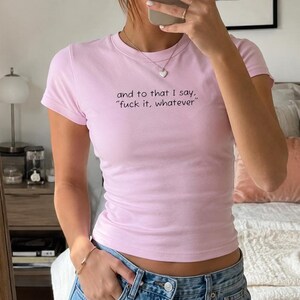 Pode incluir: Uma t-shirt de manga curta em rosa claro com o texto impresso a preto: "and to that I say, "fuck it, whatever"". A peça tem gola redonda e um corte justo, combinada com calças de ganga azuis.