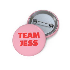Op de afbeelding: Twee ronde, roze buttons met een speldrug. Eén button toont de rode tekst "TEAM JESS". De andere button is gedeeltelijk zichtbaar en toont de zilveren speldrug. De buttons zijn waarschijnlijk voor een team of evenement.