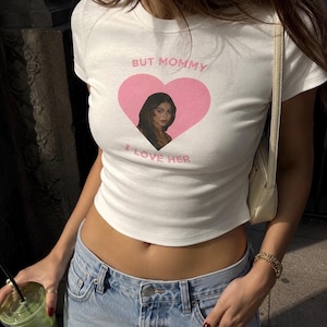 May include: A white short-sleeved crop top featuring a pink heart graphic on the chest. Inside the heart is a photographic portrait of a person with long dark hair. The text BUT MOMMY I LOVE HER is printed in pink above and below the heart.