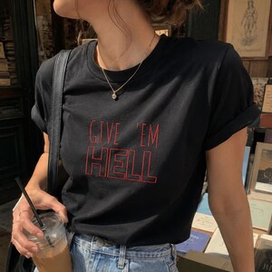 以下が含まれることがあります： 赤い文字で「GIVE 'EM HELL」と書かれた黒いTシャツ。青いジーンズと金のネックレスと合わせています。ストローの入った透明なプラスチックカップが手に持たれています。