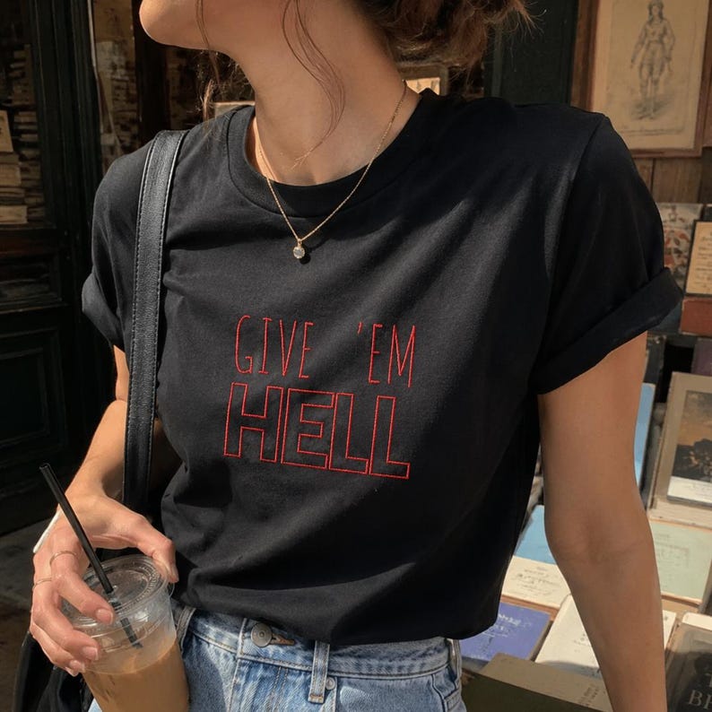 以下が含まれることがあります： 赤い文字で「GIVE 'EM HELL」と書かれた黒いTシャツ。青いジーンズと金のネックレスと合わせています。ストローの入った透明なプラスチックカップが手に持たれています。