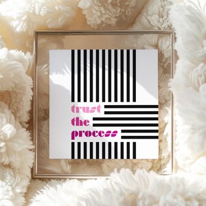 Puede incluir: Impresión artística cuadrada enmarcada con las palabras "trust the process" en rosa, sobre un fondo blanco con líneas negras verticales y horizontales. La impresión se muestra sobre una superficie blanca y esponjosa.