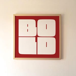 以下が含まれることがあります： 赤い背景に「BOLD」の文字が大きく白抜きで描かれたフレーム入りアートプリント。 作品は自然な木製のフレームに収められ、白い壁に掛けられています。