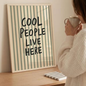 May include: A framed print with a cream background and vertical muted green stripes. The text "COOL PEOPLE LIVE HERE" is in bold black letters. A small notebook with grey stripes sits on a wooden surface.