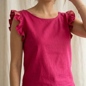 May include: A vibrant fuchsia sleeveless top featuring a wide, rounded neckline and prominent ruffled cap sleeves. The garment is crafted from a textured fabric with a visible weave, providing a structured yet relaxed fit that sits at the waistline.