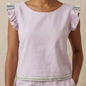 May include: A light purple sleeveless top featuring a round neckline and ruffled cap sleeves. The sleeve edges and the bottom hem are accented with a horizontal striped pattern in shades of blue, green, and white on a neutral background.