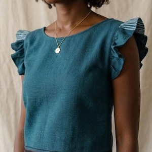 Op de afbeelding: Een blauwgroene top met korte mouwen, een ronde halslijn en ruches aan de mouwen. De mouwen hebben een subtiel wit en blauw gestreept patroon aan de onderkant. Een dunne, donkere horizontale band is zichtbaar langs de onderzoom.