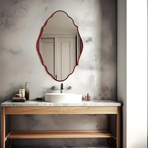 Puede incluir: Un espejo ornamental con marco rojo cuelga sobre un tocador de baño. El espejo tiene una forma ovalada estilizada con un borde festoneado. El tocador tiene una encimera de mármol, un lavabo blanco y una base de madera con un estante inferior.