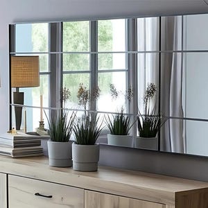 Op de afbeelding: Een rechthoekige spiegel met een rasterpatroon hangt boven een houten dressoir. De spiegel reflecteert een raam en planten. Twee lampen met beige tinten en decoratieve voetstukken staan op het dressoir, samen met potplanten.