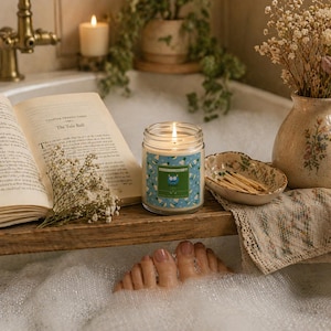 May include: A lit candle in a glass jar with a blue and green label, sitting on a wooden tray with an open book and a small dish of matches. The scene is set in a bathtub with bubbles.