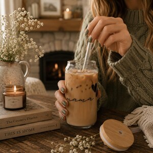 Puede incluir: Un vaso transparente en forma de lata lleno de café helado, con la palabra "mama" en escritura cursiva y un corazón. Se inserta una pajita transparente. Una tapa de madera está cerca. El vaso lo sostiene una persona con un suéter verde.