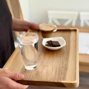 Op de afbeelding: Een houten dienblad met een glas water, een kopje koffie en een klein wit schaaltje met stukjes pure chocolade. De lade is rechthoekig met afgeronde hoeken.