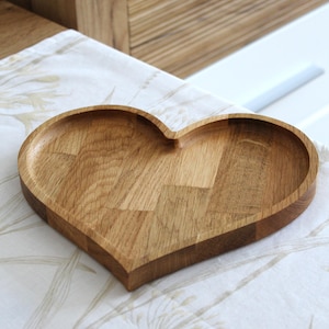 Op de afbeelding: Een hartvormig houten dienblad met een natuurlijke houtnerf afwerking. De lade is gemaakt van lichtbruin hout en heeft een licht verhoogde rand. De lade is ongeveer 25 cm breed en 25 cm lang.