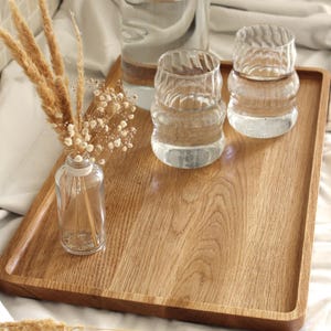 May include: A wooden serving tray holds a clear glass pitcher and two ribbed glasses, all filled with water. A small glass vase with dried flowers sits on the tray. The tray is made of light-colored wood.