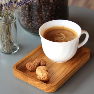 May include: A white coffee cup filled with coffee sits on a wooden tray with two cookies. A glass jar of coffee beans and a small glass jar of lavender are in the background. The scene is set on a gray table.