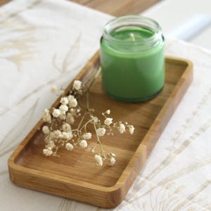 以下が含まれることがあります： 木製の長方形の小さなトレイに、ガラス瓶に入った緑色のキャンドルと白い花の小枝が置かれています。 キャンドルはトレイの中央に配置され、模様のある白い表面に置かれています。 全体的な美学は自然でミニマリストです。