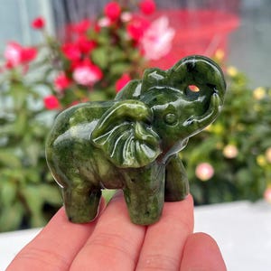 May include: A small, green jade elephant figurine. The figurine is carved with detailed features, including large ears, a trunk, and tusks. The jade has a natural, mottled appearance with variations in the green colour. The elephant is standing on four legs.