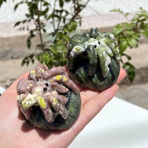 May include: Two carved stone crab figurines. One is a mix of pink, yellow, and white, while the other is green and white. Both have small black eyes and are resting on a dark green base. The crab figurines are held in a person's hand.