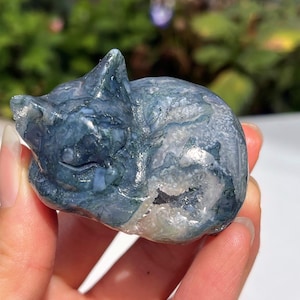 Puede incluir: Una figura de gato de piedra tallada en tonos azules, blancos y grises. El gato está acurrucado, con los ojos cerrados, y parece estar hecho de un material de piedra natural. La figura se sostiene en una mano.