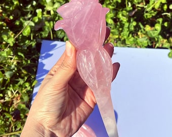 May include: A carved rose sculpture made of pink rose quartz. The rose is detailed with petals and a stem. The sculpture is held in a hand against a background of green foliage and a white surface.