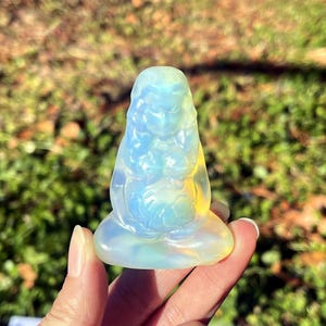 Op de afbeelding: Een doorschijnend, opalescent kristallen beeldje van een zittende figuur in een meditatieve pose. De sculptuur heeft een lichtblauw en geel kleurenschema, met details die een sereen gezicht en vloeiend haar suggereren. Het beeldje wordt in een hand gehouden tegen een vage groene achtergrond.
