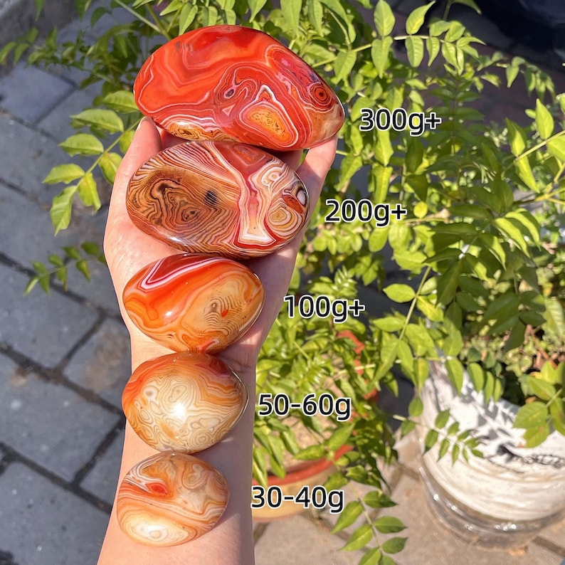 Peut inclure: Cinq pierres d'agate polies, lisses et arrondies, aux nuances orange, rouge et blanc avec des motifs en bandes. Les pierres sont &eacute;tiquet&eacute;es avec des poids : 300g+, 200g+, 100g+, 50-60g et 30-40g, indiquant leurs tailles respectives.