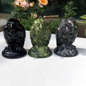 Könnte beinhalten: Drei geschnitzte Eulenfiguren aus Stein in Schwarz, Grün und Grau. Die Eulen sitzen auf runden Sockeln und haben detaillierte Federstrukturen. Der Hintergrund zeigt grünes Laub und rosa und orange Rosen.