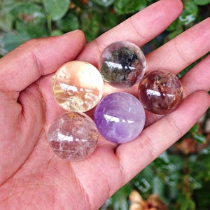 May include: Five polished crystal spheres in various colours, including clear, amber, purple, and brown. The spheres are held in a hand, with a blurred green background. The crystals are round and appear semi-transparent.