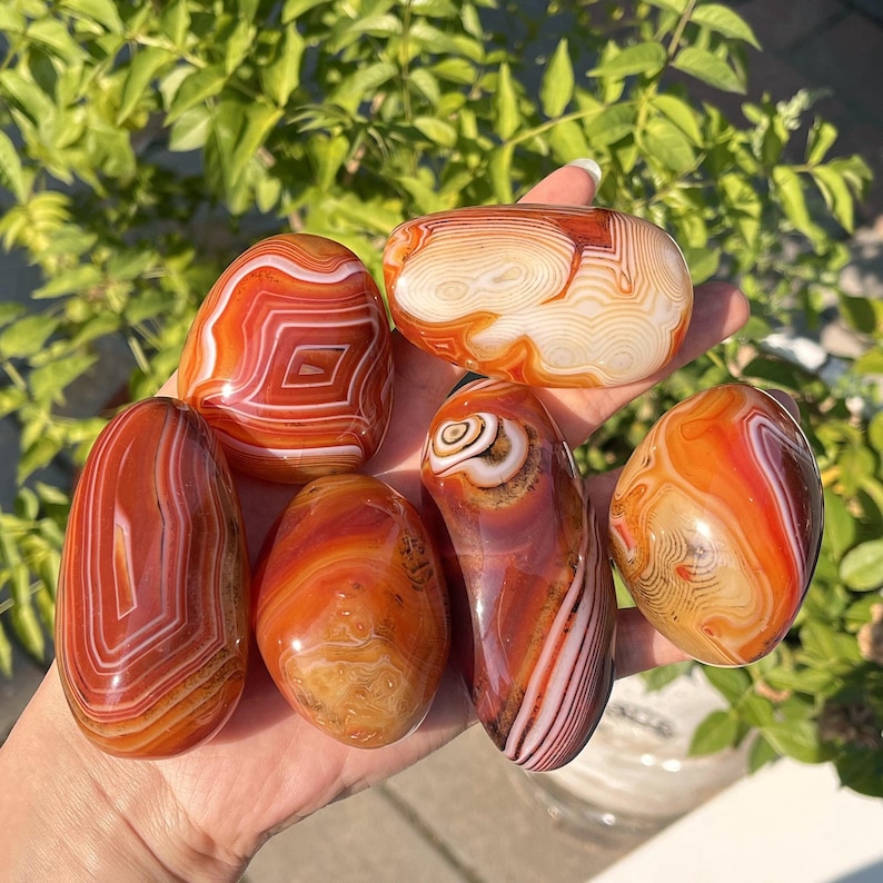 Peut inclure: Un ensemble de six pierres polies, lisses et arrondies, pr&eacute;sentant des bandes vibrantes orange, rouges et blanches. Chaque pierre affiche des motifs concentriques complexes et des couches d'agate, avec une texture de surface brillante.