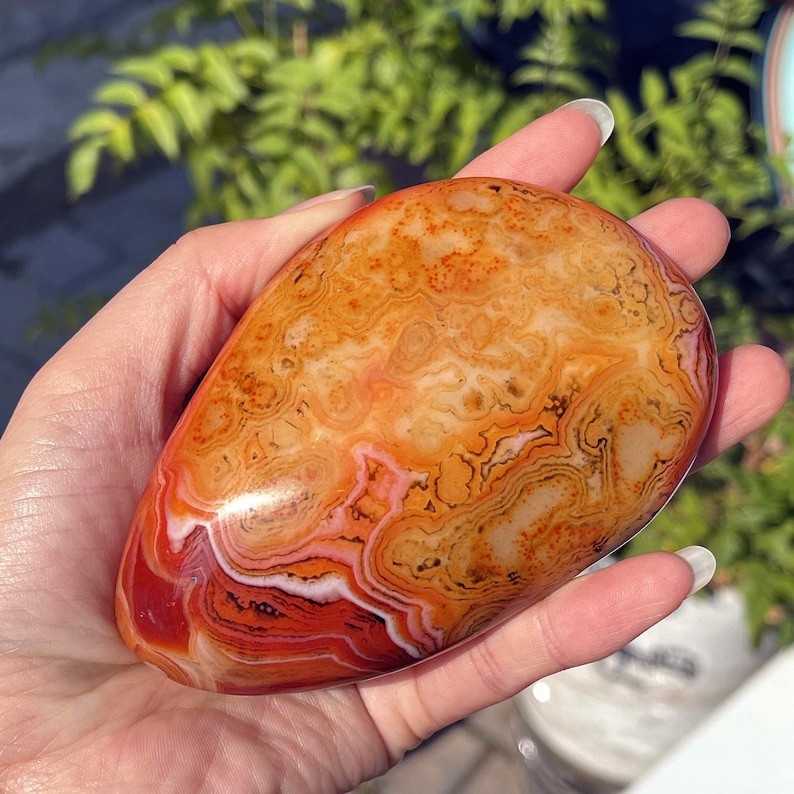 Peut inclure: Une pierre d'agate polie et lisse en forme de goutte, pr&eacute;sentant des bandes complexes et tourbillonnantes de couleurs orange vif, rouge profond, blanc cr&egrave;me et brun terreux. La surface offre une finition brillante et r&eacute;fl&eacute;chissante.