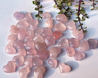 May include: A collection of polished rose quartz heart-shaped stones in varying shades of pink. The stones are scattered on a white surface, with green leafy vines in the background. The hearts are smooth and translucent.