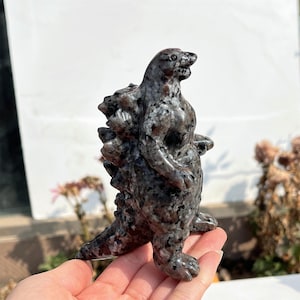 May include: A detailed sculpture of a Godzilla-like creature carved from speckled grey stone. The figure stands upright with a textured back and a detailed face. The sculpture is held in a hand, showcasing its size and craftsmanship.