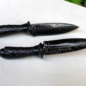 Puede incluir: Dos dagas decorativas negras con intrincadas tallas florales en los mangos y las hojas. Las dagas tienen un acabado brillante y una punta. Los mangos son redondeados y texturizados.