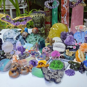 May include: A collection of various gemstone sculptures and crystals. The assortment includes a yellow skull, a green Buddha statue, amethyst clusters, and carved animal figures. The colors range from purple and green to yellow and orange, with a variety of shapes and sizes.