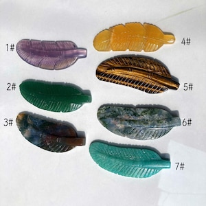Può includere: Sette piume di pietre preziose scolpite in vari colori. Le piume sono numerate da 1 a 7. I colori includono viola, verde, giallo, marrone e verde-blu. Ogni piuma ha venature dettagliate e una finitura liscia. Le piume sono disposte su una superficie bianca.