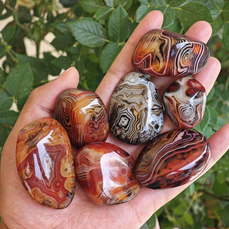 Peut inclure: Six pierres d'agate polies, lisses et arrondies, pr&eacute;sentant des motifs de bandes concentriques complexes dans des tons de rouge profond, orange br&ucirc;l&eacute;, cr&egrave;me, blanc et brun fonc&eacute;. Chaque pierre affiche des couches min&eacute;rales naturelles.