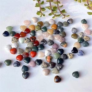 May include: A selection of heart-shaped stones in a variety of colours, such as red, green, pink, and black. The stones are arranged on a white surface, with some green foliage in the background. These polished stones are suitable for crafts or as decorative accents.